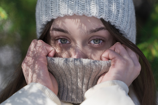 Cropped shot teenager girl pulling collar of knitted sweater up to his eyes, hiding face from frosty winter weather and cold temperature looking at camera. Model 17-year-old with gray eyes, no make-up in knit hat, down jacket. Part of series of photographs