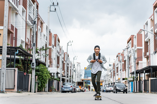 Asian young male ride surf skate board on street on the way to work. Attractive extreme athletic guy feel happy and relax enjoy activity in free time play skate for health and wellbeing on the road.
