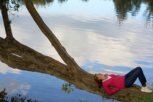 A woman lying thoughtful and relaxed on a fallen tree over the river listening to the sounds of nature. Concept: connection with nature and outdoor lifestyle.
