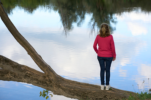 Person rear view in pink sweater standing on a fallen tree in front of the river with the reflection of the forest in the background. Concept: connection with nature and outdoor lifestyle