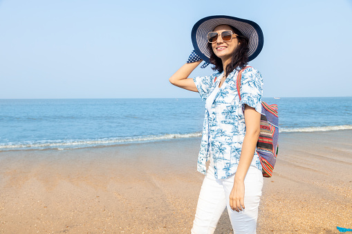 Beautiful young indian girl with travel bag and hat, sunglasses enjoying vacation, holiday at beach.