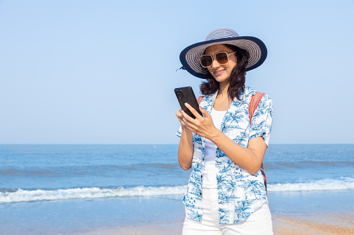 young Indian girl in summer casual clothes using mobile smart phone at beach. Travel, vacation concept.