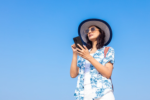 young Indian girl in summer casual clothes using mobile, smart phone at beach. Travel, vacation concept.