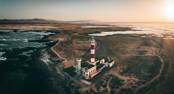 Tostón Lighthouse or El Cotillo Lighthouse in moody sunset light. The lighthouse is situated on the northwestern coast of Fuerteventura Island close to the village of El Cotillo. The lighthouse was opened in 1897, and consisted of a 7 m high plain masonry tower on one corner of the single storey keeper's house. Aerial Drone View over the surrounding coast at sunset. Stitched Panorama. Tostón Lighthouse, El Cotillo Lighthouse, El Cotillo, La Olivia, Fuerteventura Island, Canary Islands, Spain - Africa