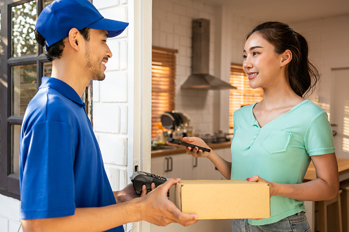 Asian young delivery man delivering package to female customer at home. Attractive postman in blue uniform working, deliver a box of fruit and vegetable groceries to woman infront of the door in house