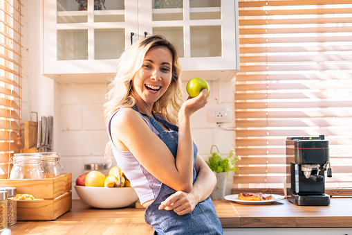Caucasian young woman holding an apple on hands in the kitchen at home. Attractive beautiful girl wearing apron feeling happy and relax, enjoy cooking healthy fruit for lose weight and diet in house.