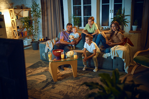 Smiling parents and children sitting on sofa in living room in the evening and watching together TV. Comedy movie or show. Concept of family, leisure time, relaxation, childhood and parenthood
