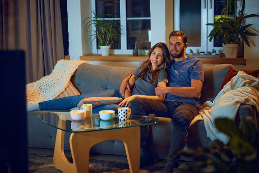 Loving young couple, man and woman sitting on sofa in evening at home,, hugging, cuddling and watching tv together. Concept of family, leisure time, relaxation, childhood and parenthood