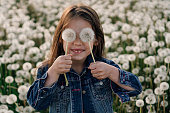 Joyful girl standing in field of white dandelions and holding two flowers in hands covering eyes with fluffy blowballs.