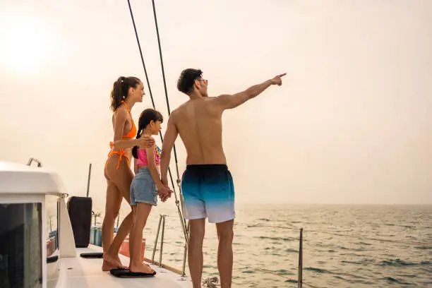 Photo of Caucasian happy family walking on deck of yacht while yachting outdoor. Young beautiful couple hanging out and spend time with daughter while catamaran boat sailing during holiday summer trip together
