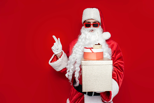 santa claus in christmas costume and glasses holds boxes with gifts and shows with his hand to the side on copy space on red background
