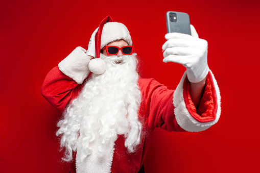 santa claus in hat and festive glasses uses smartphone and takes selfie on red background, man in santa costume talks via video link online