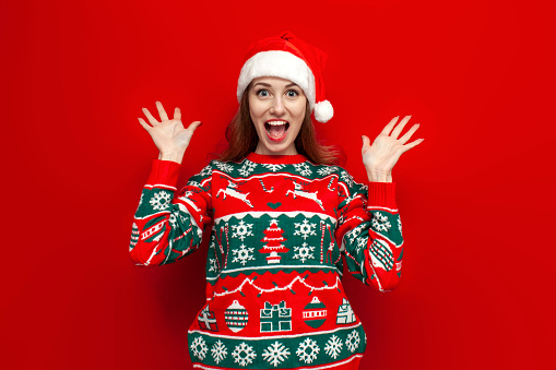 young cute girl in christmas sweater and santa hat rejoices and shouts on red background, woman in christmas clothes raises her hands and shows her palms