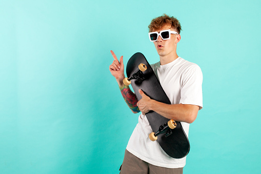 young cheerful tattooed guy in white t-shirt holds skateboard and smiles on blue isolated background, hipster skater in sunglasses shows his hand to the side and advertises copy space