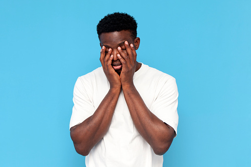 tired african american man in white t-shirt in depression covers his face with his hands on blue isolated background, sleepy indifferent man is bored and unhappy