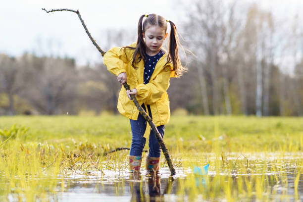 istock Girl holding branch while standing in puddle and looking down with focused eyes, preparing for game with paper boats. 1796249739