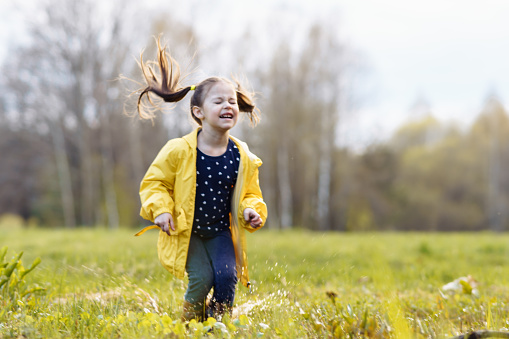 Gorgeous child in yellow jacket and rubber boots squinting eyes and jumping on puddle, playing with water on green site after rain. Little girl with ponytails splashing pool and enjoying leisure.