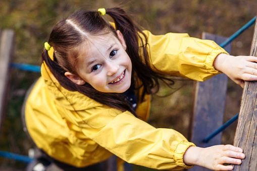 View from above of adorable little girl with two ponytails, adorned in bright yellow jacket, climbing on staircase constructed at playground and looking at camera with charming and shiny smile.