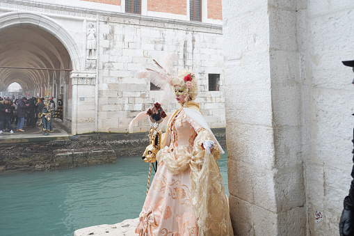 Venice, Italy - February 16 2023: Fully masked person standing by a canal near old building with stone walls in Venice, time of traditional Carnival in winter