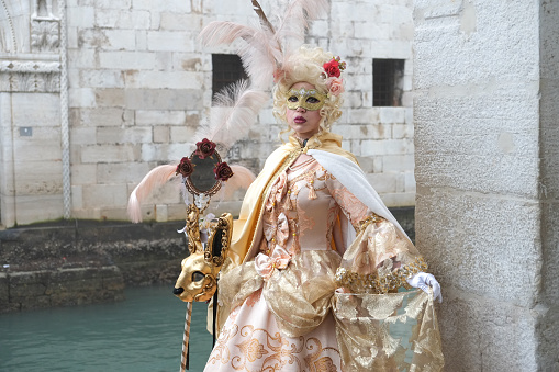 Venice, Italy - February 16 2023: Fully masked person standing by a canal near old building with stone walls in Venice and rehearsing for traditional Carnival in winter