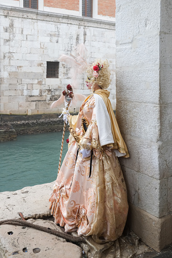 Venice, Italy - February 16 2023: Fully disguised person standing by a canal near stone wall of an old building in Venice, time of traditional Carnival in winter