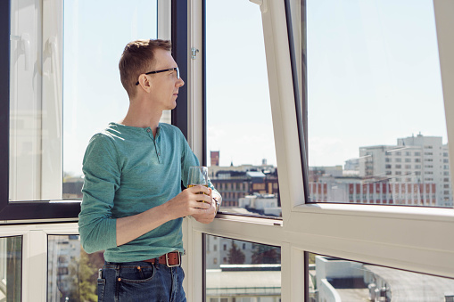 Positive emotions. Man in turquoise T-shirt with long sleeves stands on balcony of house, apartment, office with glass of juice and smiles, looking at camera. City and buildings. Tourism and travel.