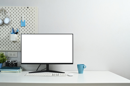 Creative workplace with blank computer monitor, coffee cup, houseplant, stationery and peg board on wall.