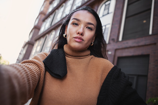 Candid Self portrait of brunette woman looking at camera outdoors. Beautiful female fashion model on street of city takes selfie photo.