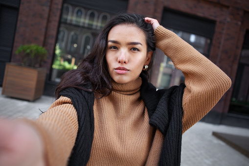 Selfie portrait of brunette Asian woman seriously looking at camera outdoors. Beautiful female fashion model on street of city takes headshot.