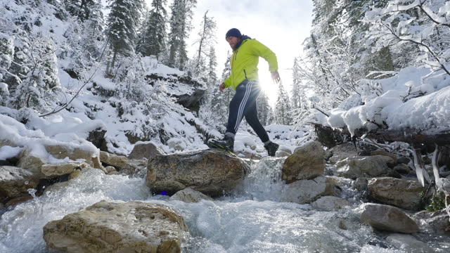 SLO MO Young Man Crossing River Flowing Through Rocks in Snow-Covered Coniferous Trees in Dolomites