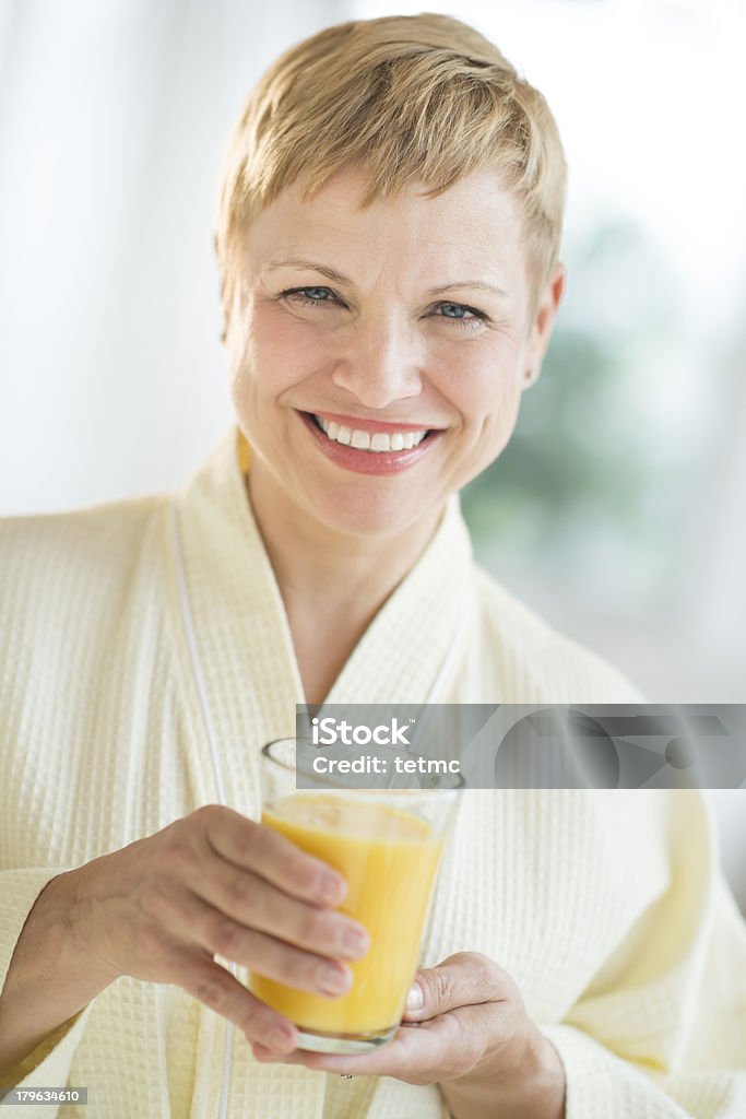 Happy Woman Holding Glass Of Juice Portrait of happy mature woman in bathrobe holding glass of juice at home Drinking Glass Stock Photo