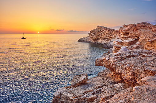 Wide-angle view of the cove of Punta Galera and its peculiar sandstone formations jutting over the sea, located on the western coast of Ibiza in the surroundings of the town of Sant Antoni de Portmany (also known as San Antonio Abad), at sunset. A sailing boat crossing calm waters reflecting the golden light of a Mediterranean summer sunset, picturesque clouds, colourful cliffs. High level of detail, natural rendition, realistic feel. Developed from RAW.