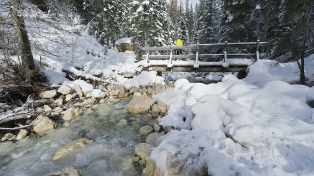 SLO MO Male Hiker Walking on Wooden Bridge Over Flowing River in Snow Covered Forest in Dolomites