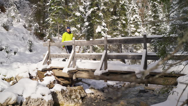 SLO MO Young Male Hiker Walking on Snow Covered Wooden Bridge Over Flowing River in Forest, Dolomites