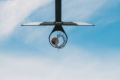 A basketball ball is entering in the basketball hoop. View from below, sky in the background.