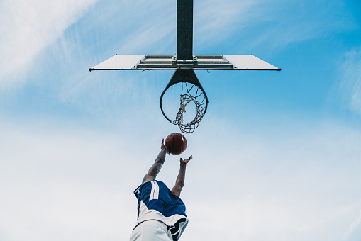 Low angle view of a young adult man scoring a goal in a basketball court. Sky in the background.