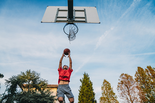 Low angle view of a young adult man scoring a goal in a basketball court. Sky and trees in the background.