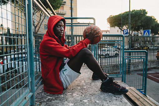 Portrait of a young adult man while he's sitting on the basketball court's bleachers. He's wearing a red hooded sweatshirt. He's looking at camera.