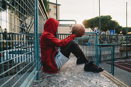 A young adult man is resting on the basketball court's bleachers. He's wearing a red hooded sweatshirt. Sunset time.