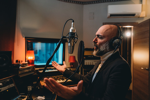 Singer is recording music in a professional recording studio. He's singing nad he's making gestures with hands.