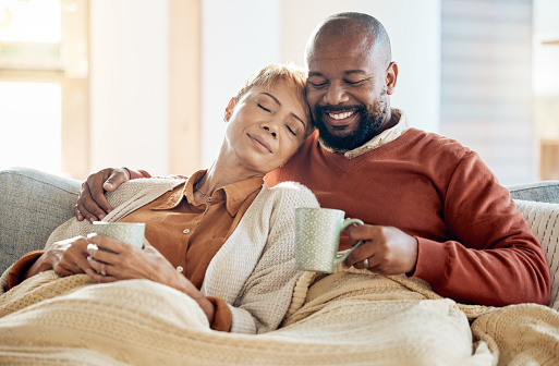 Couple relax on sofa with coffee, blanket and smile on winter weekend morning in home. Peace, comfort and love, happy black man and tired woman, cozy time on couch with drink in living room together.