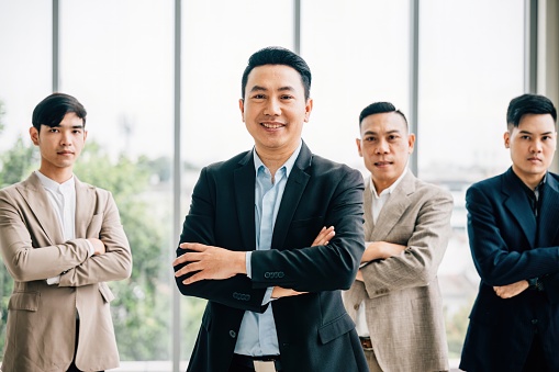 A confident businessman in smart casual attire smiles, arms crossed in a creative office. Diverse Asian male and female colleagues stand together at a startup, showcasing teamwork and success.