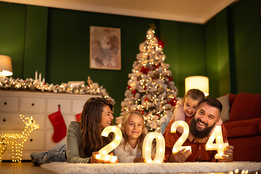 Parents and children having fun celebrating New Year together at home, lying by the Christmas tree holding illuminative numbers 2024