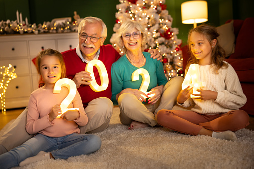 Grandparents celebrating New Years Eve at home with kids, sitting by the Christmas tree, holding illuminative numbers 2024 representing the upcoming New Year