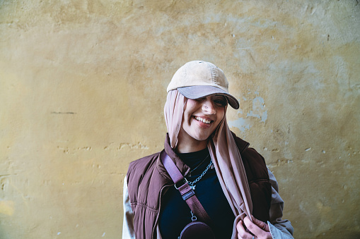 Portrait of a young adult woman against a yellow background. She's wearing a fashionable hijab with an hat.