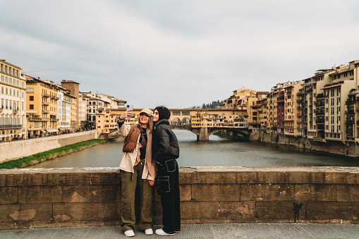 Two friends are taking a selfie on Santa Trinita bridge with Ponte Vecchio in the background. They are wearing fashionable clothes.