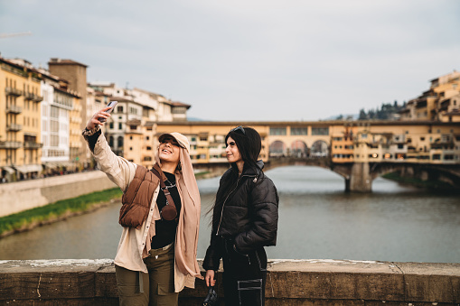 Two friends are taking a selfie on Santa Trinita bridge with Ponte Vecchio in the background. They are wearing fashionable clothes.