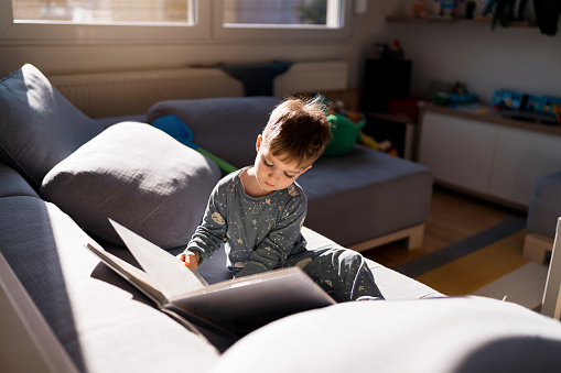 While sitting on a sofa in pajamas, cute Caucasian toddler boy reading a child book.
