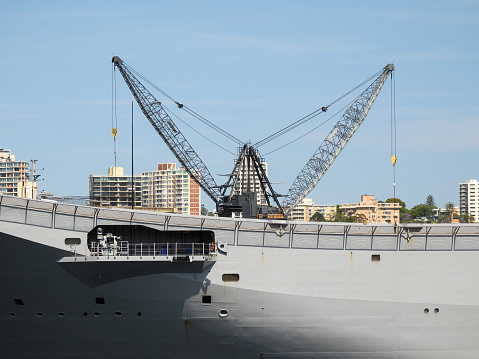 The bow and flight deck of HMAS Adelaide of the Royal Australian Navy docked at Garden Island, Sydney Harbour.  In the background are two dockside cranes. In the distance are residential buildings of Elizabeth Bay.  This image was taken on a sunny afternoon on 11 November 2023.