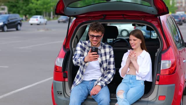 Happy man and woman looking at phone screen while making selfie. Couple sitting on the trunk car. Transportation, technology, trip, travel and lifestyle concept. Real time